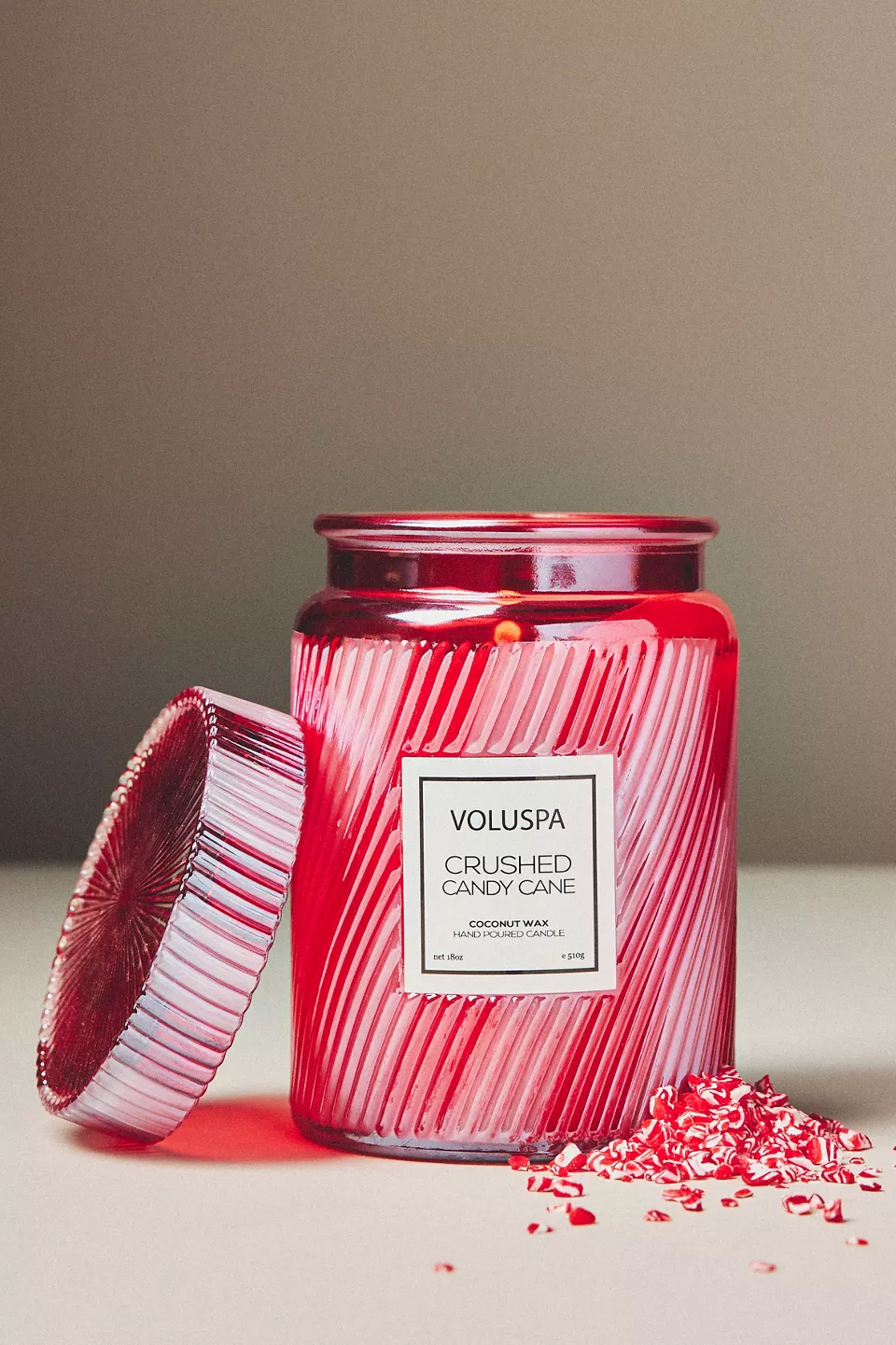 A candle in a red jar with the lid off and crushed candy canes in front of it