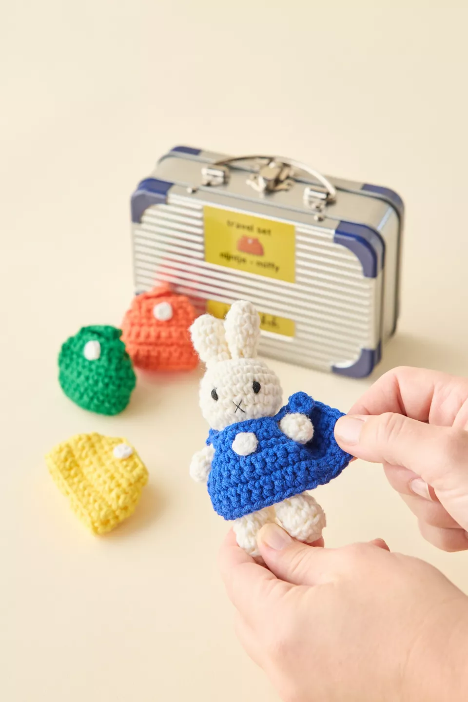A silver briefcase next to three small crochet sweaters and two hands holding a white crochet bunny in a blue sweater