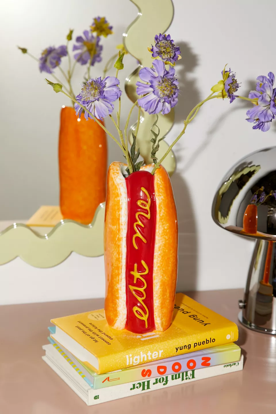 A vase shaped like a hot dog on top of a stack of three books with purple flowers inside